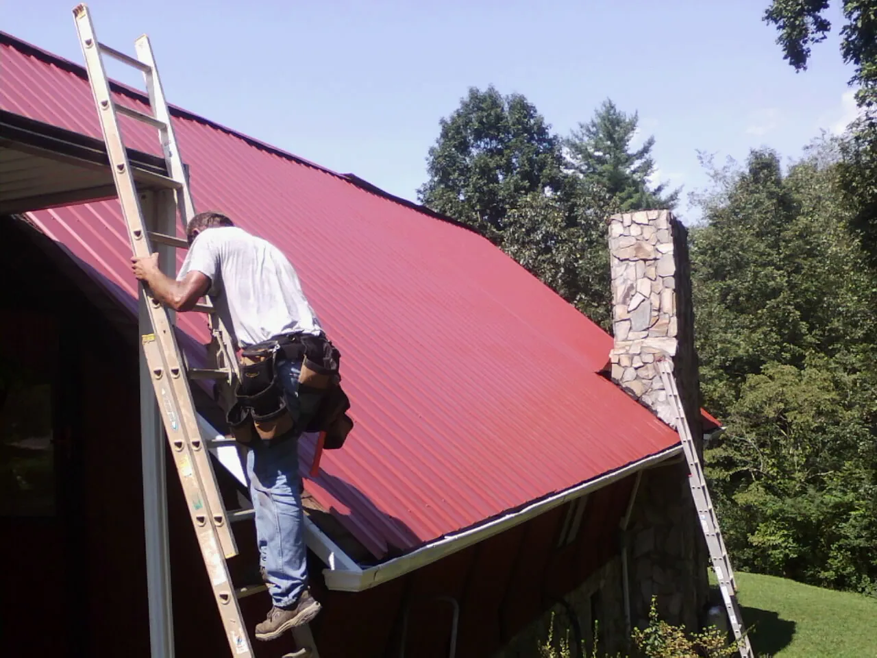 Roofer on a ladder beside a red metal roof and stone chimney in De Soto